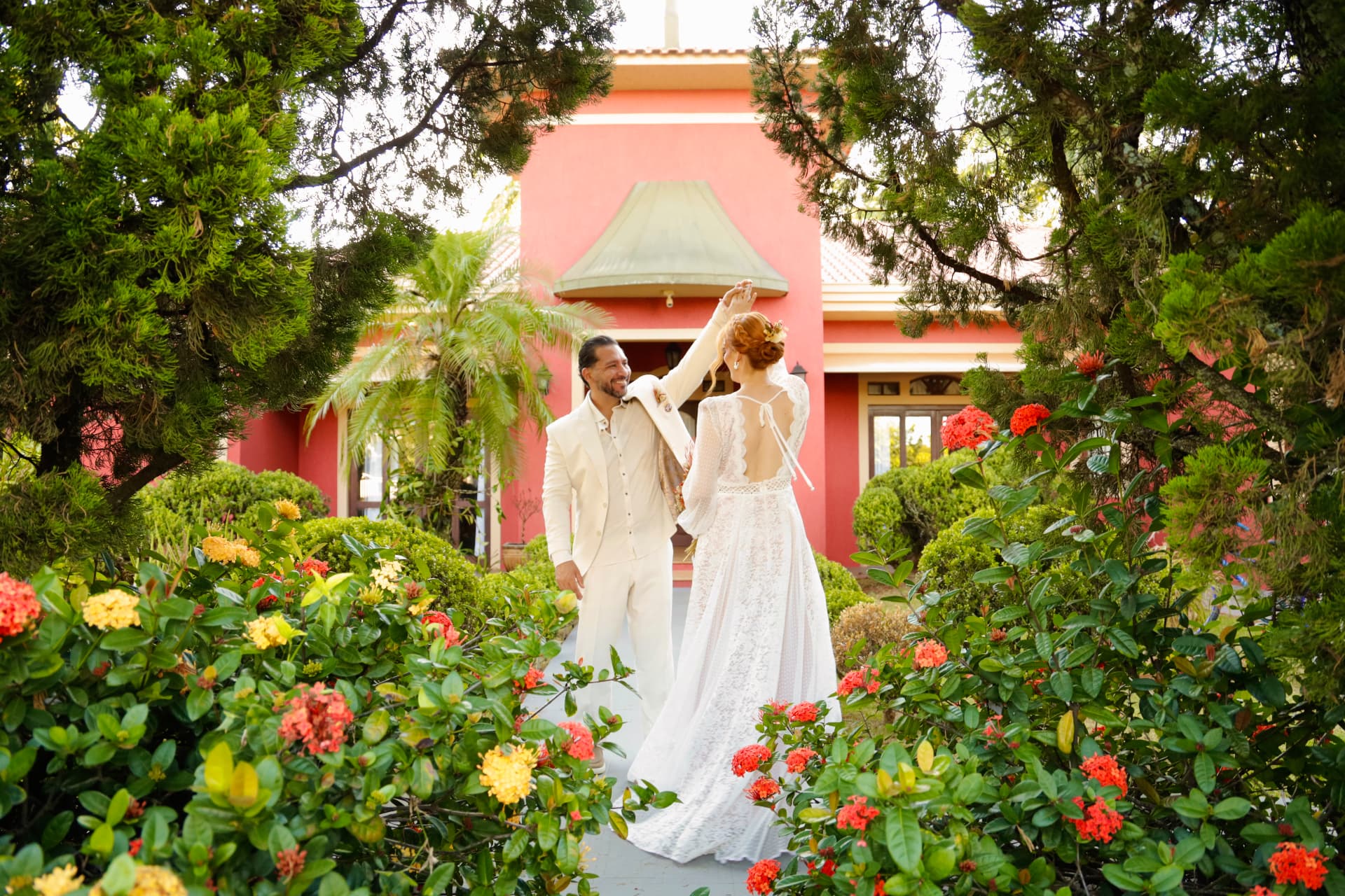 Noivos dançando em um jardim florido com uma casa cor-de-rosa ao fundo